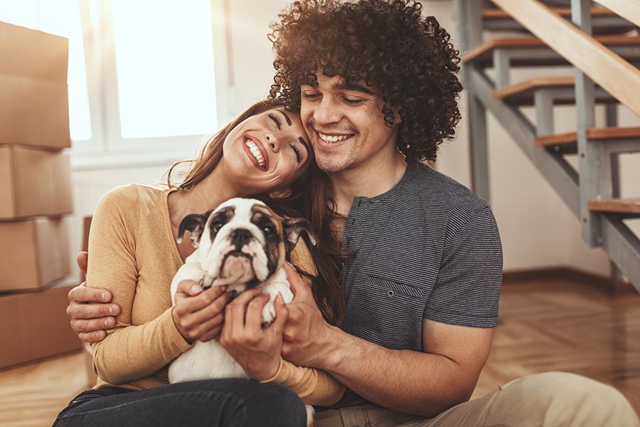 About Our Agency - Closeup View of a Cheerful Young Couple Hugging Their Dog as They Sit on the Floor in Their New Apartment