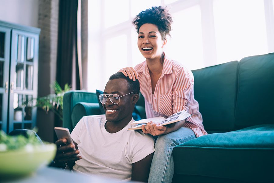 Personal Insurance - Cheerful Young Married Couple Hanging Out Together in the Living Room While Sitting on the Sofa as the Wife Holds a Photo Album and the Husband Uses His Phone